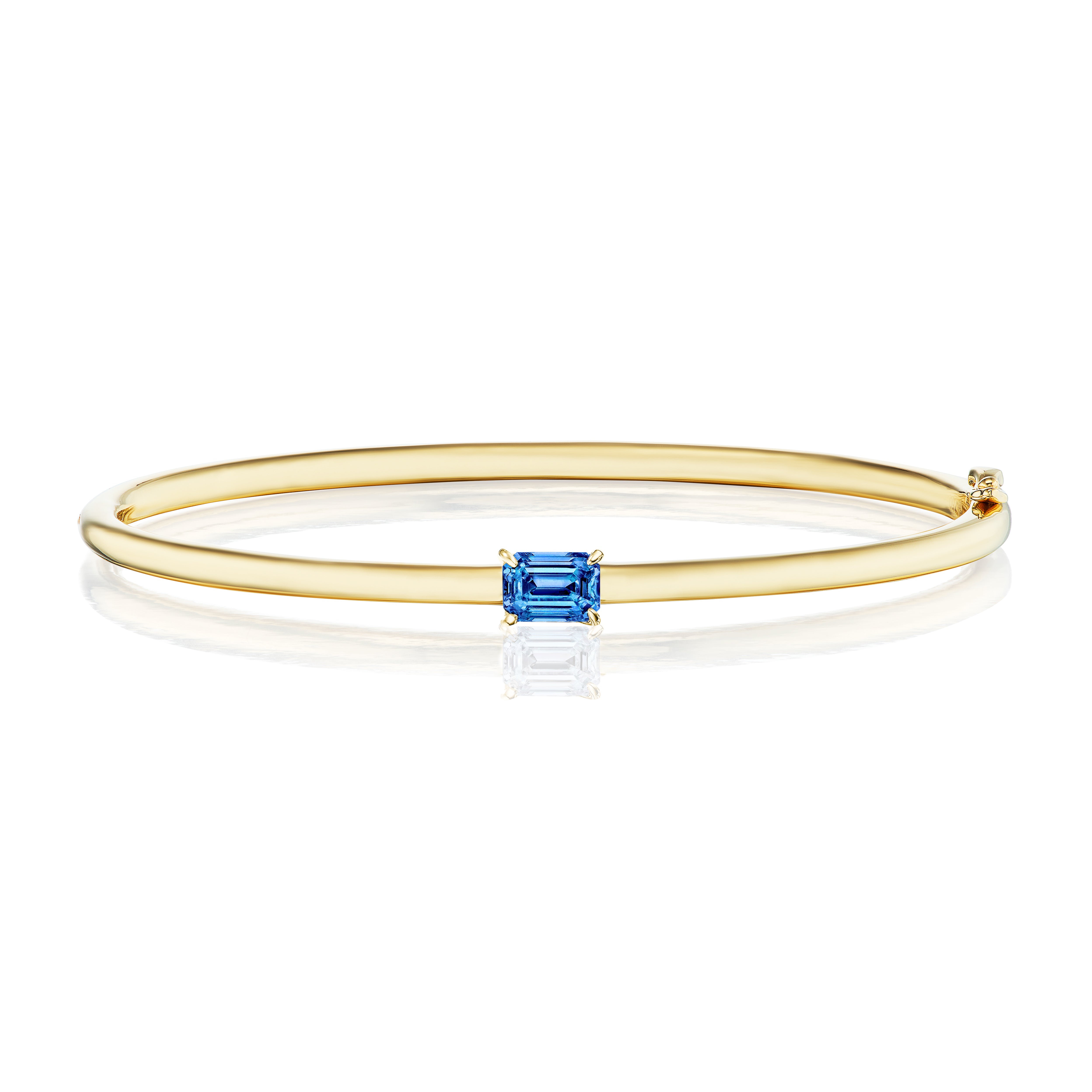 The East-West Emerald Cut Solitaire Blue Sapphire Bangle in Yellow Gold showcases a striking blue sapphire at its center, elegantly displayed against a white background.