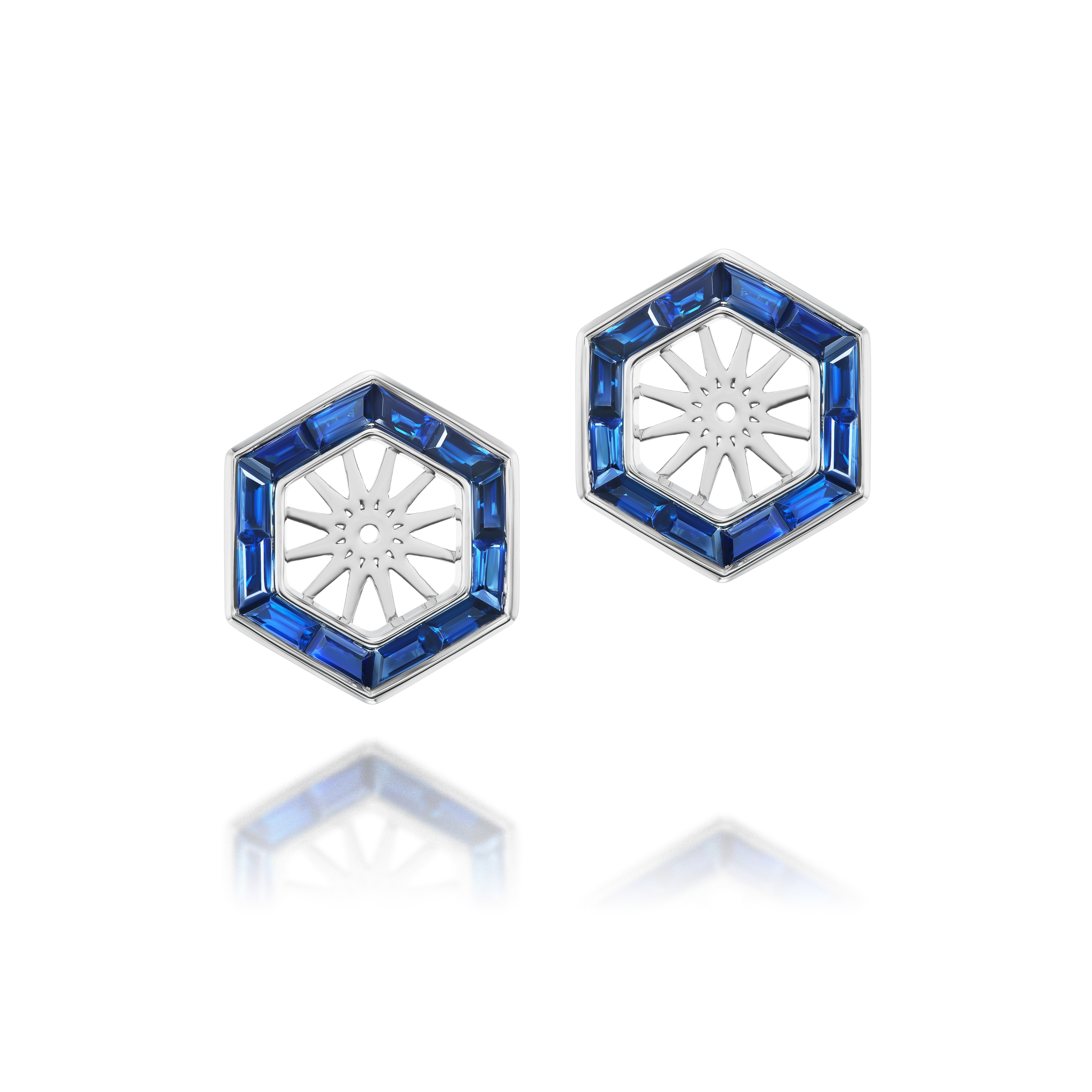 The Hexagonal Trapezoid Sapphire Stud Ear Jackets feature silver hexagon earrings with blue sapphire accents outlining the shape and a sunburst design at the center, shown on a white background with a gentle reflection.