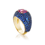 The Cobblestone Bombé Ring in Yellow Gold features a large hot pink oval sapphire and smaller round blue sapphires set in 18k gold, creating a vibrant and eye-catching cocktail ring.