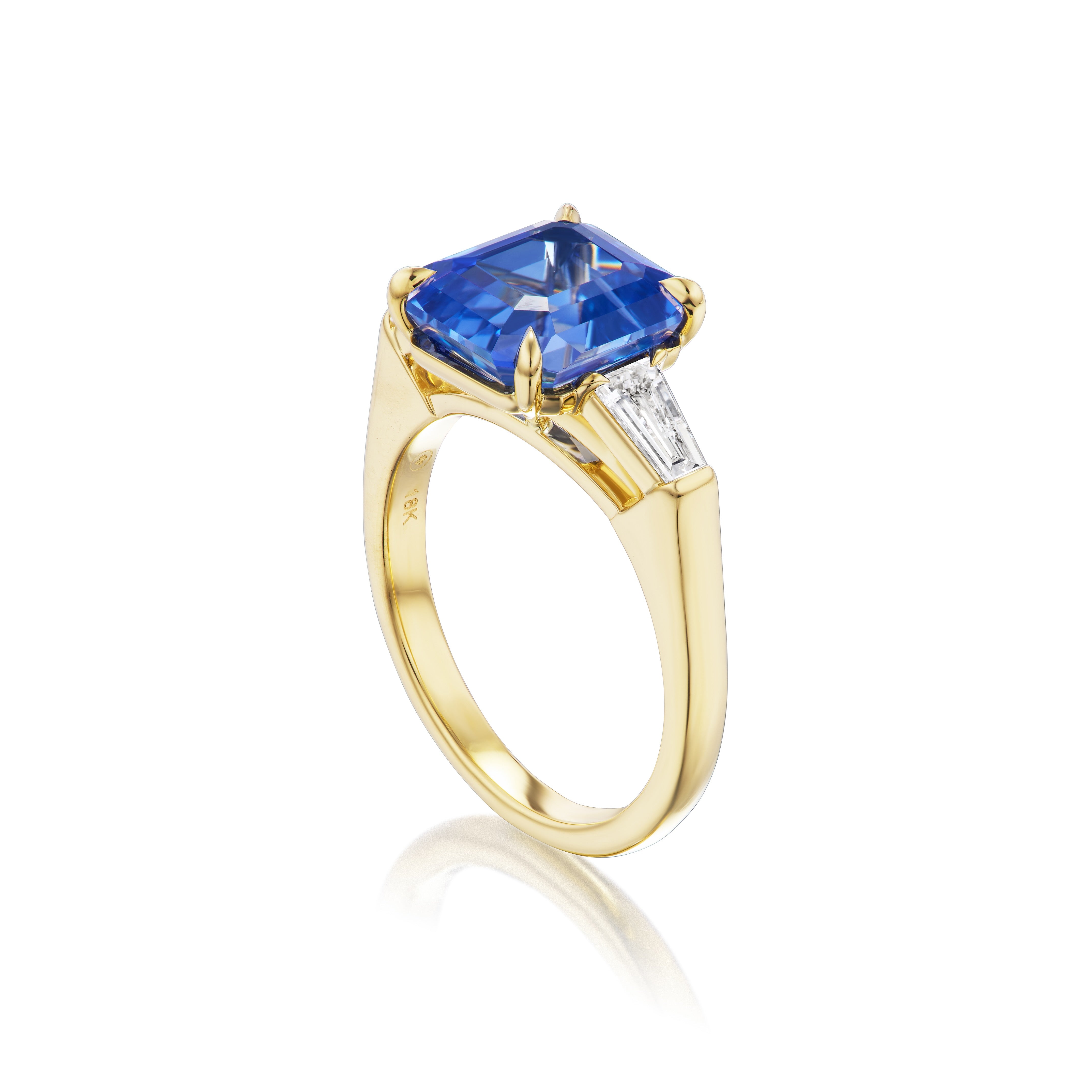 The Asscher Cut Sapphire Engagement Ring features a large Asscher cut sapphire center, accented by tapered baguette diamond side stones, all set in 18K yellow gold and shown on a white background.