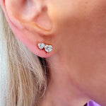 Close-up of an ear with light skin and blonde hair wearing Duo Studs with Heart Diamonds—two heart-shaped diamond earrings, one larger than the other, crafted in 18k White and Rose Gold.