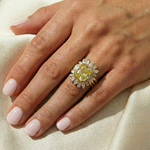A hand with light pink manicured nails rests on cream fabric, wearing the Bezel Set Cushion Cut Yellow Diamond Ring Jacket with pear and emerald cut side stones, sparkling with white diamonds on a gold band.