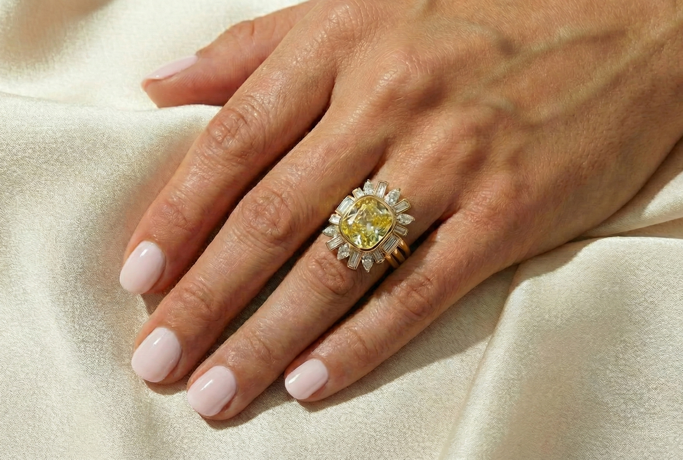 A hand with light pink manicured nails rests on cream fabric, wearing the Bezel Set Cushion Cut Yellow Diamond Ring Jacket with pear and emerald cut side stones, sparkling with white diamonds on a gold band.