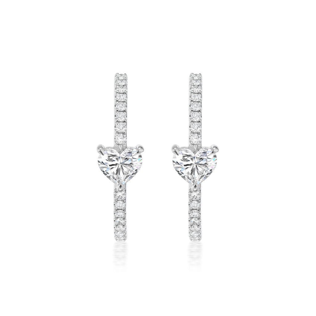 The Heart Diamond Pavé Hoops, crafted in 18K white gold, feature a heart-shaped clear gemstone at the center and are encrusted with small round diamonds, shown against a white background.