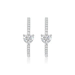 The Heart Diamond Pavé Hoops, crafted in 18K white gold, feature a heart-shaped clear gemstone at the center and are encrusted with small round diamonds, shown against a white background.