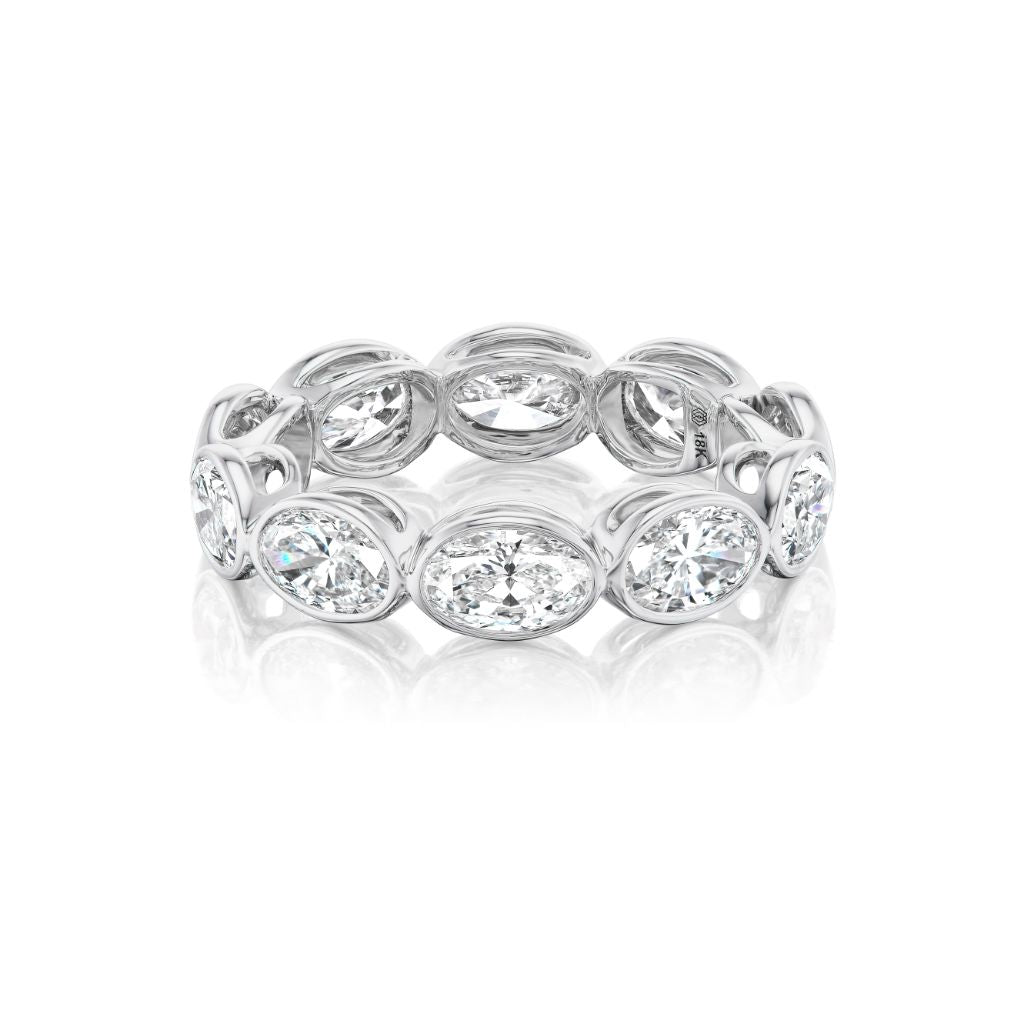 The East-West Bezel Set Oval Diamond Eternity Band in White Gold is shown on a white background with a subtle reflection, highlighting its bezel-set oval diamonds.