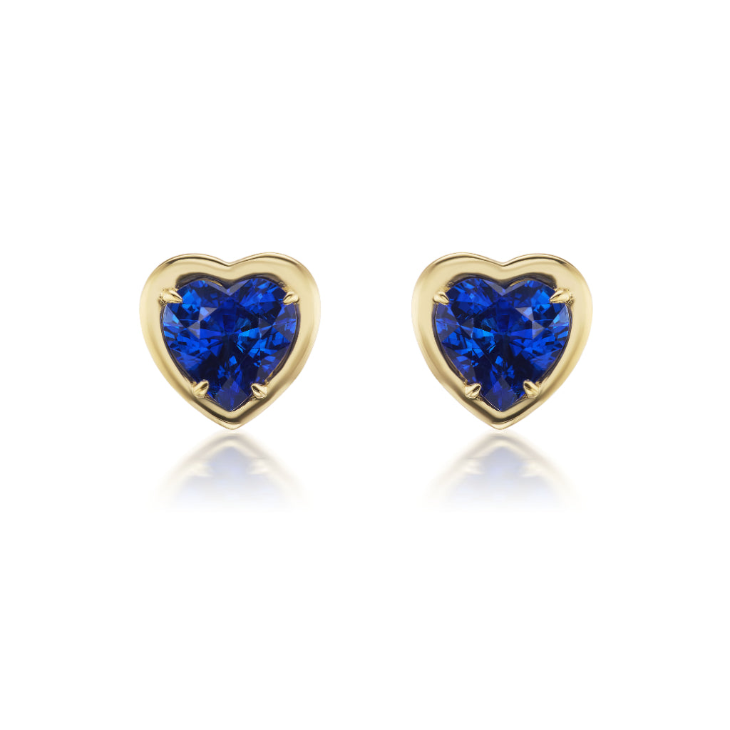 The Sapphire Heart Studs in Yellow Gold feature vivid blue heart-shaped sapphires set at the center of elegant gold heart studs, shown against a white background.