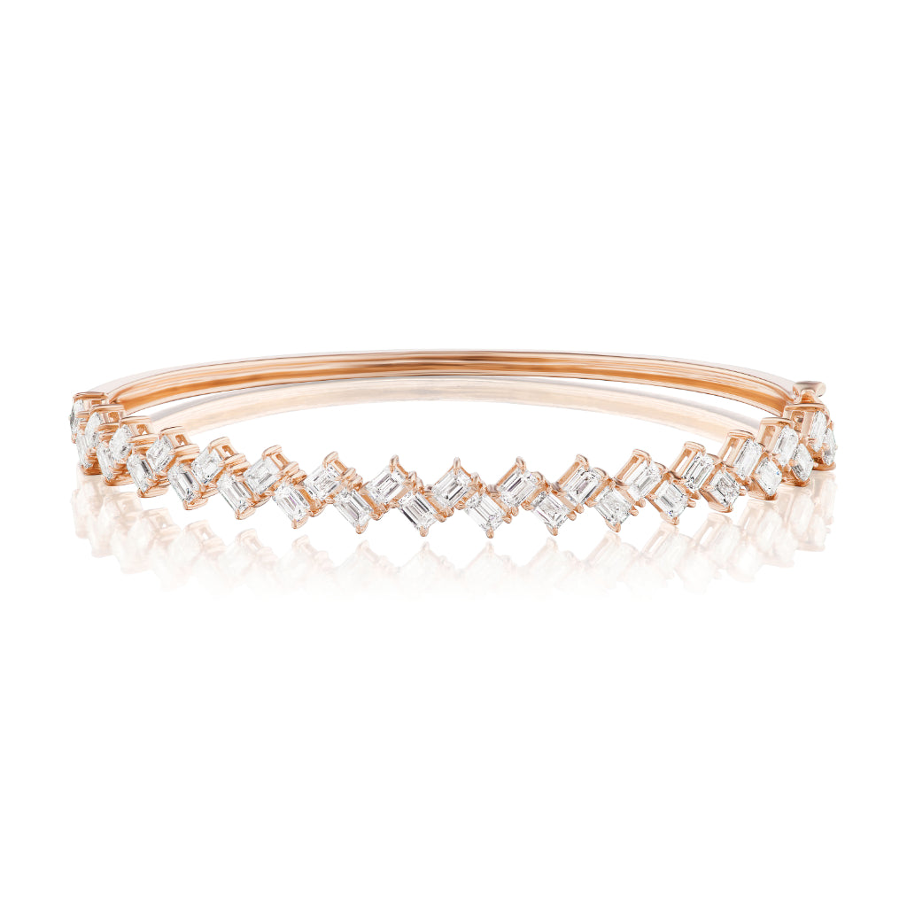 The Zig-Zag Emerald Cut Diamond Bangle, in rose gold with rectangular and square clear stones, is displayed on a reflective white background.