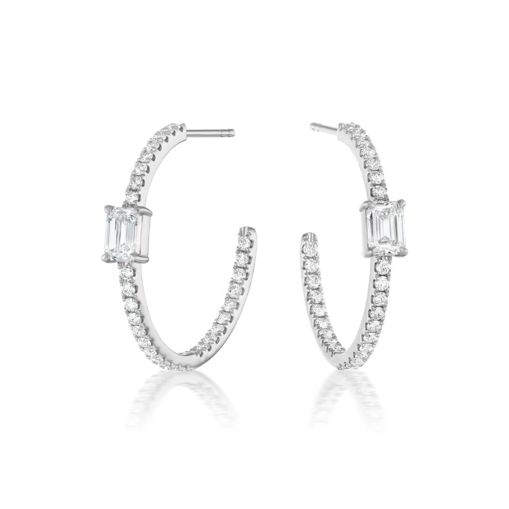 The Pavé Hoop in White Gold with Emerald Cut Diamonds features 18K white gold hoops encrusted with small round diamonds and a central emerald cut diamond, shown on a white background.