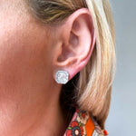 Close-up of a woman's ear wearing Mosaic Studs with Asscher Cut Diamonds in white gold. She has blonde hair and a colorful floral top. The background is softly blurred.