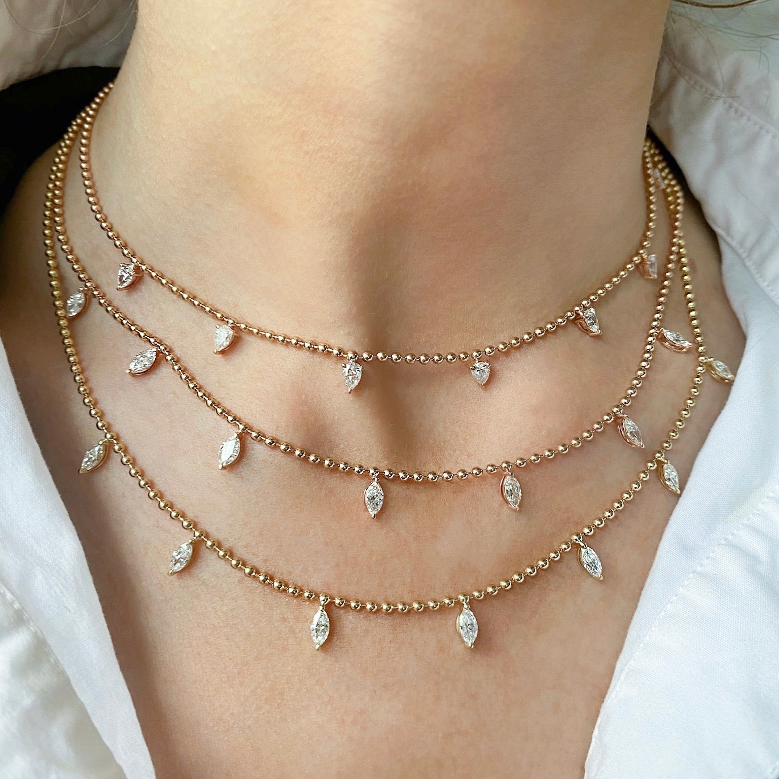 A close-up of a person wearing the Spike Ball Chain Necklace with Pear Diamonds, featuring three layered 18K gold bead necklaces with pear-shaped diamond charms, styled over a white collared shirt.