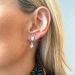 Close-up of a woman's ear wearing Drop Earrings featuring fancy light pink and white diamonds in rose gold and platinum, with a geometric design reminiscent of shield cut stones. She has straight blonde hair and wears a patterned top.